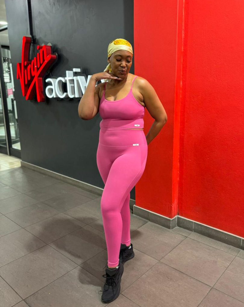 An African lady wearing fuchsia pink high-waisted leggings and strappy top standing infront of Virgin Active gym wall