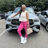 Lifestyle shot of a woman in high-waisted pink leggings and a white sleeveless crop top, white sneakers and white socks leaning against a luxury SUV and holding a beige handbag. Optimized for Premium Athleisure Streetwear South Africa.