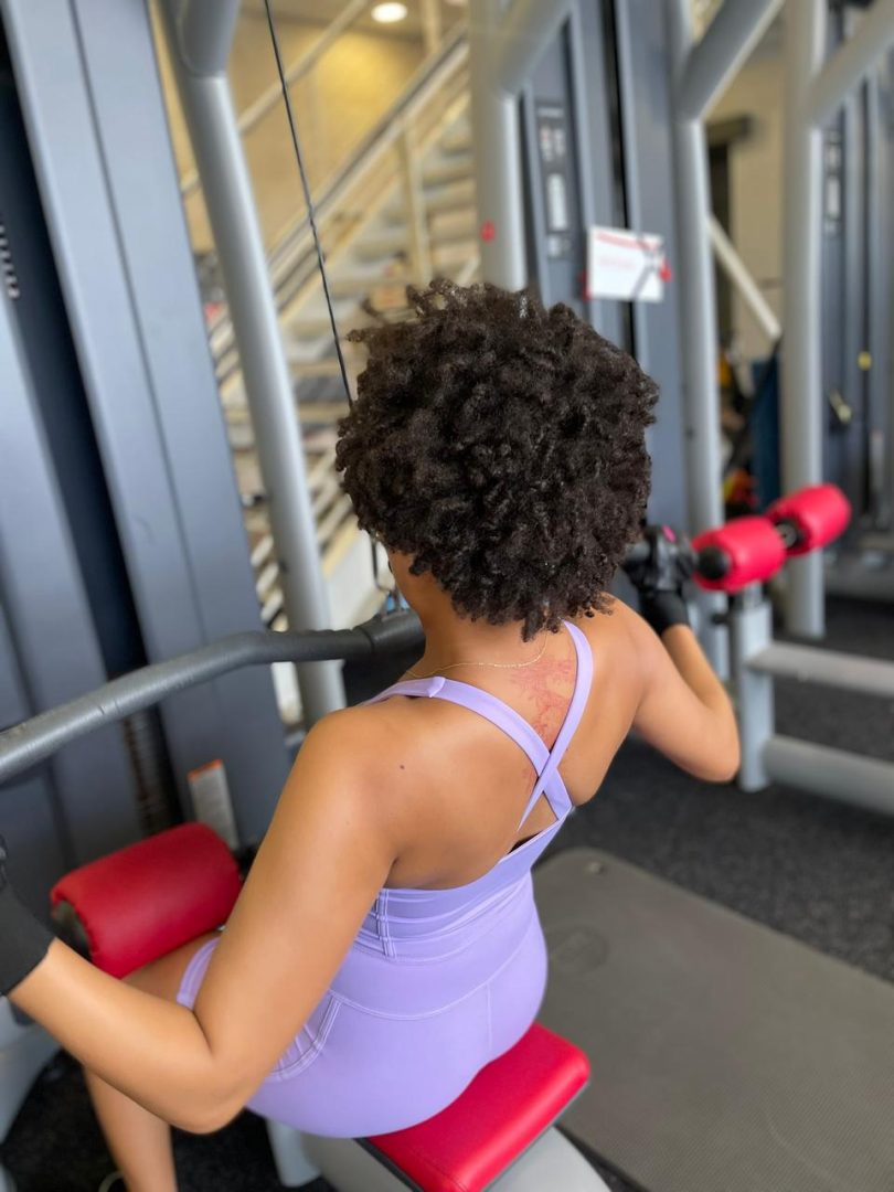 Back view of woman wearing lavender open back athleisure bodysuit using gym machine South Africa
